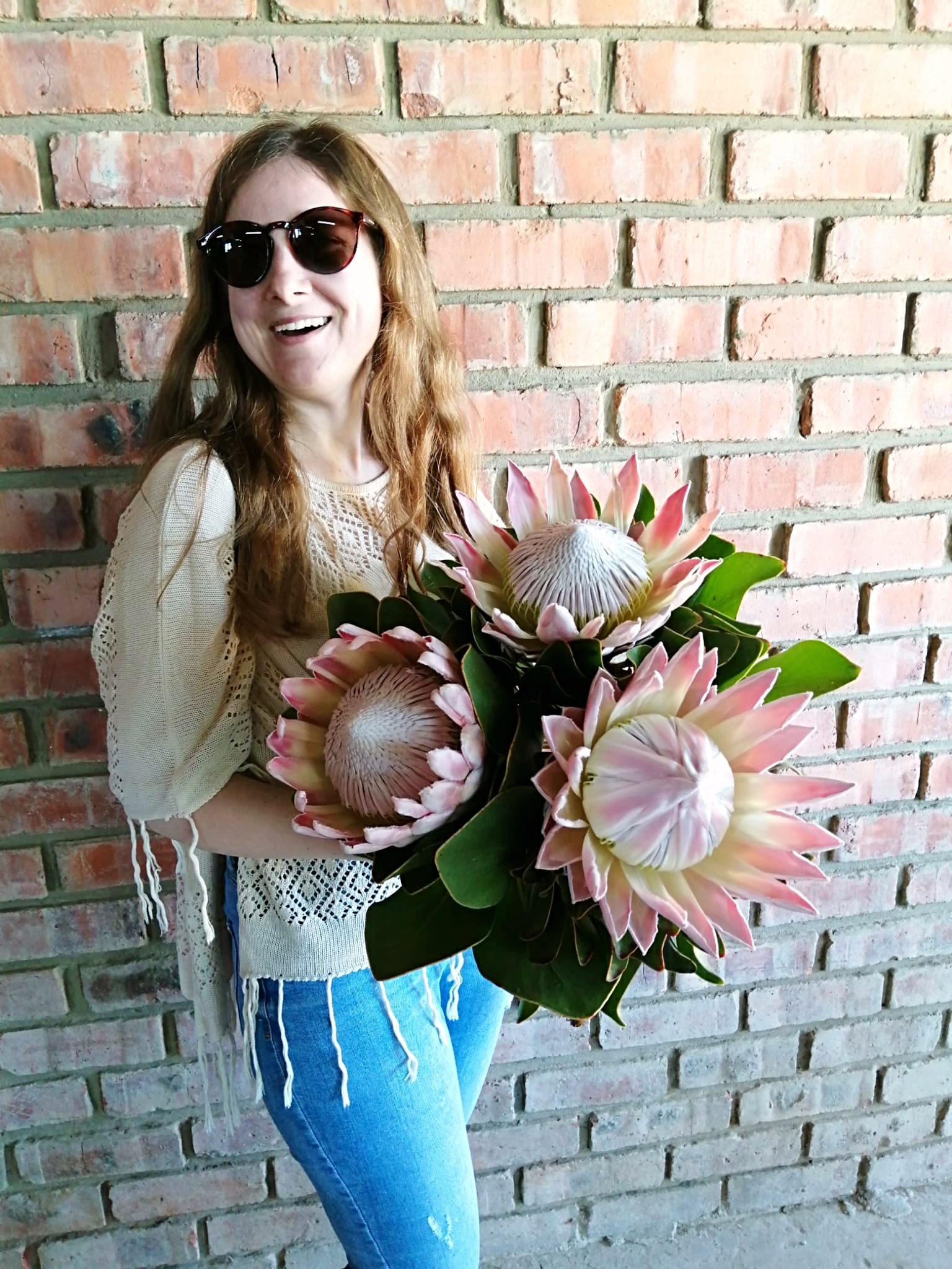 Larissa Wolfram-Kuhl mit Sonnenbrille hält große rosa Protea-Blumen vor Ziegelwand