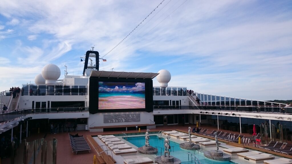 Kreuzfahrtdeck mit riesigem TV, Liegen und Pool.