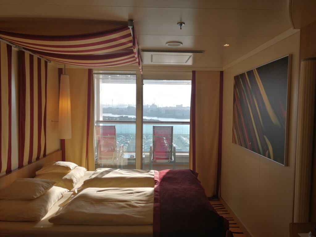 Kreuzfahrtkabine mit Bett und Blick zum Balkon mit Stühlen.