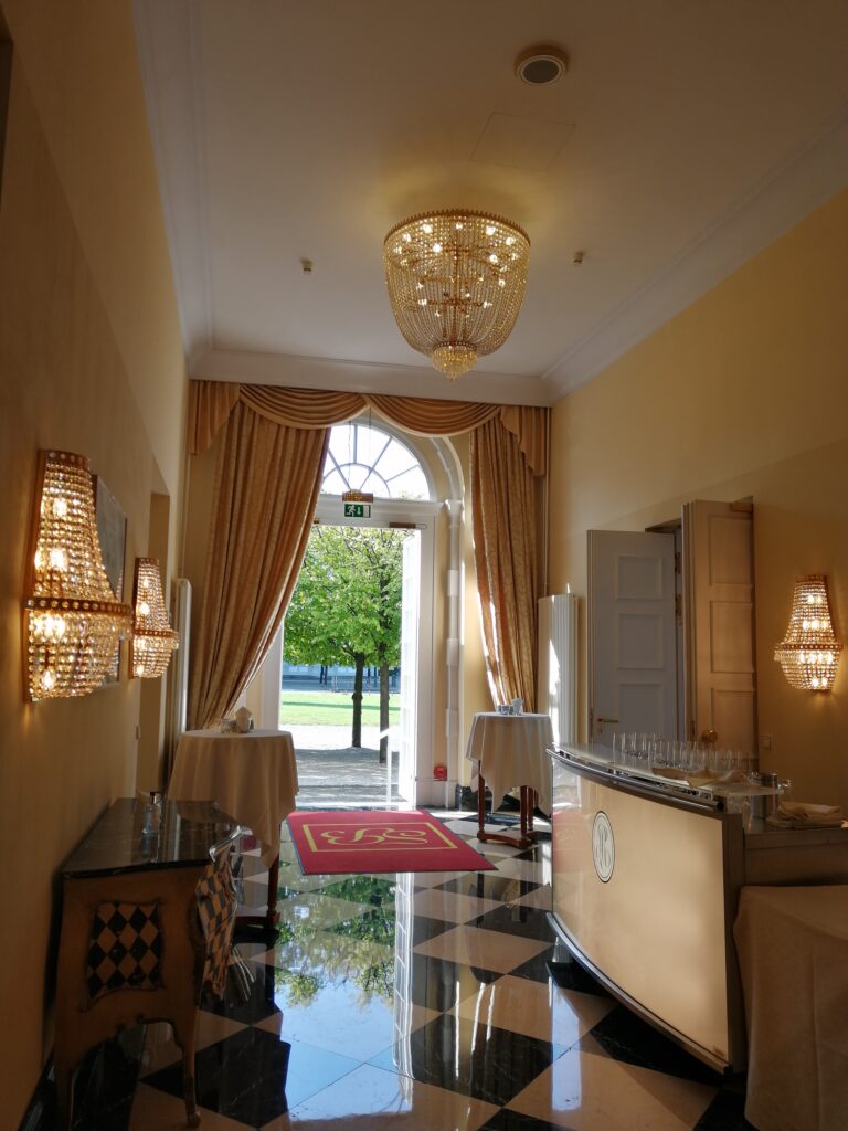 Hotelhalle mit Kronleuchter, Flur edel, Bar auf der rechten Seite und offene Tür zum Vorgarten im Hintergrund